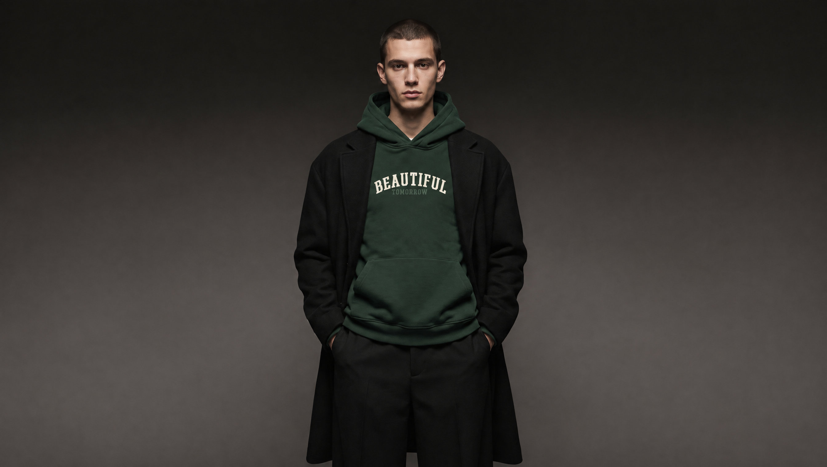 Model in the forest green Varsity Hoodie under an open black overcoat